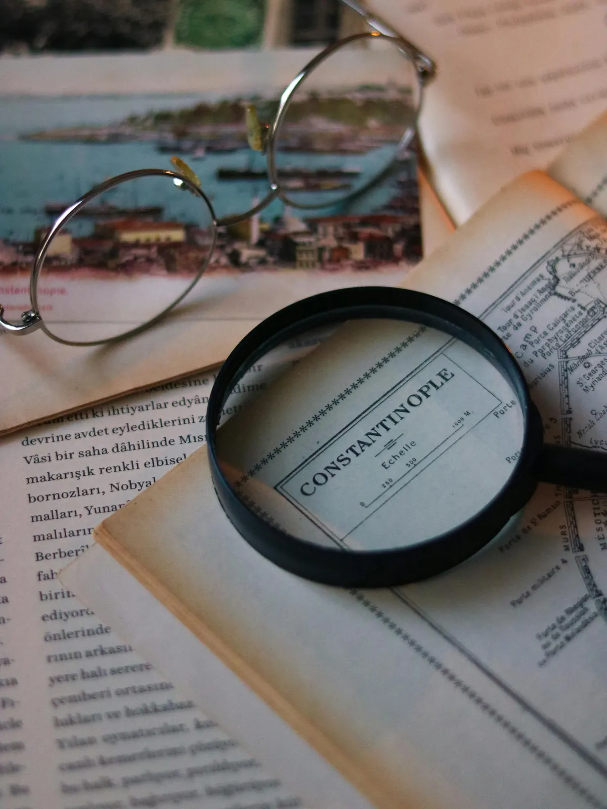 A magnifying glass on an old map of Constantinople, beside wire-rimmed glasses and a tinted postcard